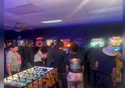 A group of people plays arcade games and foosball in a dim, neon-lit arcade room booming with colorful screens.