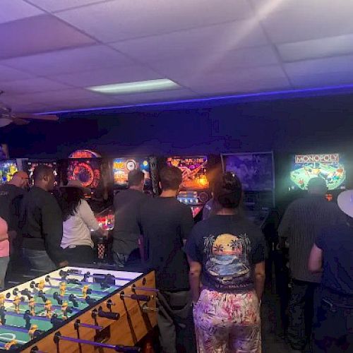 A group of people plays arcade games and foosball in a dim, neon-lit arcade room booming with colorful screens.