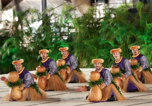 A group of small wooden figurines dressed in grass skirts and purple tops dance around coconuts on a tiled floor.