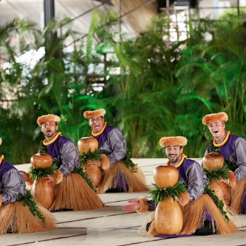 A group of small wooden figurines dressed in grass skirts and purple tops dance around coconuts on a tiled floor.