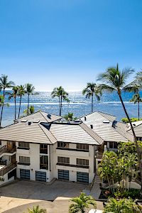 A sunny coastal scene with a row of beige townhomes, palm trees, and a sparkling ocean backdrop on a clear day.