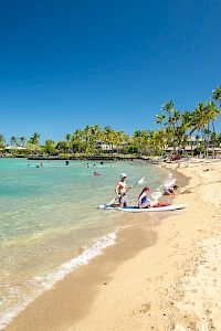 A sunny tropical beach with clear turquoise water, golden sand, palm trees, and people swimming and relaxing near the shore.