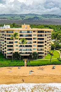 A beachfront hotel with beige apartment building facing a sandy shore, palm trees, a pool, and green lawns; ocean waves roll in beyond.