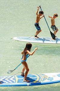 Three people stand on two paddleboards in calm water: two boys on a high-stand paddleboard, a girl on a lower board, all paddling and balancing as they move.