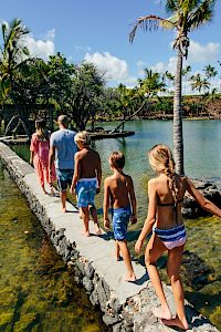 A group of people walk hand-in-hand along a narrow stone path over a clear lagoon, palm trees and blue skies in a tropical setting.