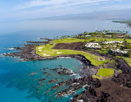 A coastal golf course with green fairways set on rocky lava shores, blue ocean, and distant islands under a clear sky.