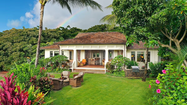 A tropical-style villa with a lush garden, outdoor seating, and a covered patio, set against trees and vibrant landscaping.