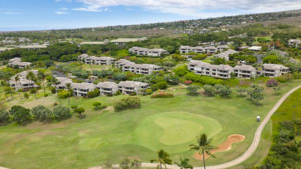 Aerial view of a coastal resort with white villas, green lawns, palm trees, and a winding path surrounding a small golf course.