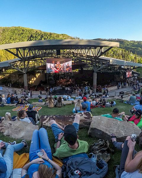Outdoor concert in a hillside park; a large stage with a crowd lounging on grass and rocks, trees and hills in the background.