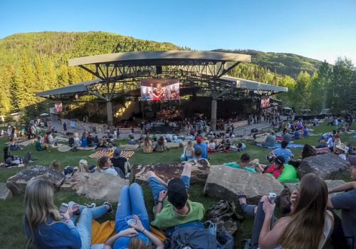 Outdoor concert in a hillside park; a large stage with a crowd lounging on grass and rocks, trees and hills in the background.
