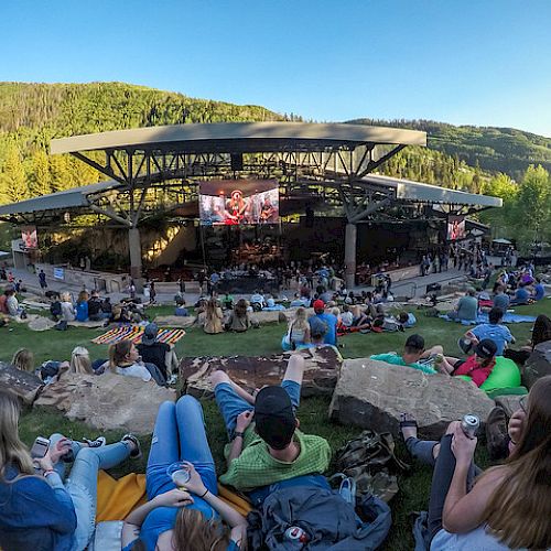 Outdoor concert in a hillside park; a large stage with a crowd lounging on grass and rocks, trees and hills in the background.