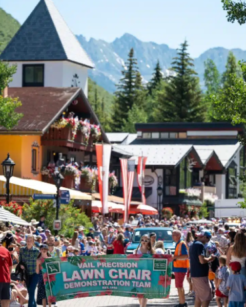 A crowded outdoor festival with a banner reading &ldquo;Law Chair Demonstration Team,&rdquo; colorful buildings, tents, and a mountain backdrop with cheering attendees.