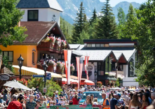 A crowded outdoor festival with a banner reading &ldquo;Law Chair Demonstration Team,&rdquo; colorful buildings, tents, and a mountain backdrop with cheering attendees.