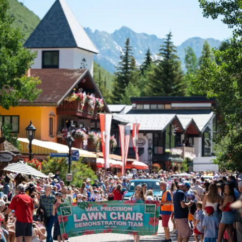 A crowded outdoor festival with a banner reading &ldquo;Law Chair Demonstration Team,&rdquo; colorful buildings, tents, and a mountain backdrop with cheering attendees.