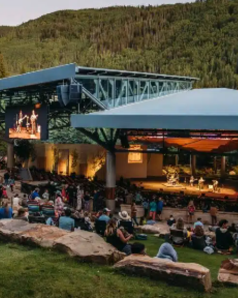 Outdoor amphitheater at dusk with a crowd seated on benches, a stage with screens, and surrounding green hills.