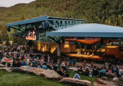 Outdoor amphitheater at dusk with a crowd seated on benches, a stage with screens, and surrounding green hills.