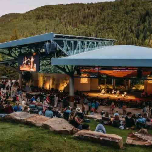 Outdoor amphitheater at dusk with a crowd seated on benches, a stage with screens, and surrounding green hills.