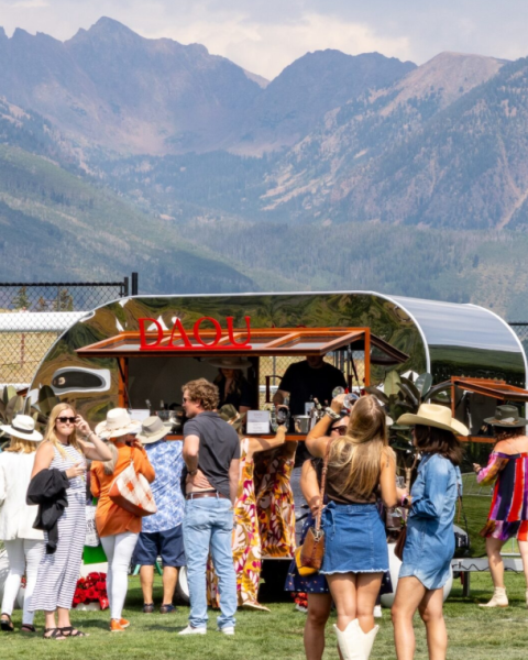 A sunny outdoor festival at a mountain venue with a line of people ordering drinks from a curved bar; attendees wear casual, summery outfits.