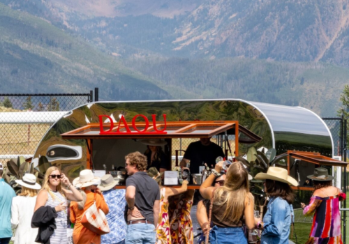 A sunny outdoor festival at a mountain venue with a line of people ordering drinks from a curved bar; attendees wear casual, summery outfits.