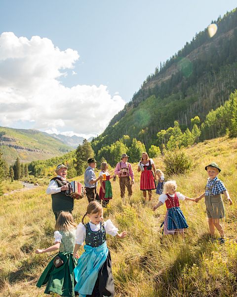 A group of children in traditional Bavarian clothes play on a sunny hillside among wildflowers, with mountains and trees in the background.
