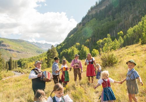 A group of children in traditional Bavarian clothes play on a sunny hillside among wildflowers, with mountains and trees in the background.