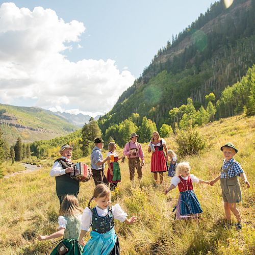 A group of children in traditional Bavarian clothes play on a sunny hillside among wildflowers, with mountains and trees in the background.