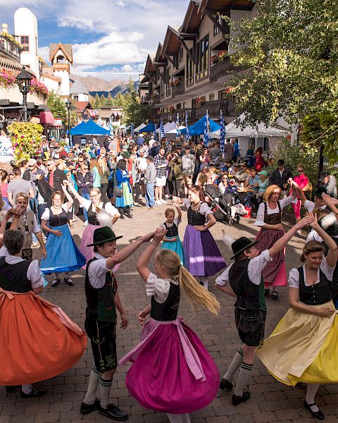 A festive street scene with people dancing in colorful skirts, vendors, and crowds lining a sunny cobblestone avenue&mdash;spirited celebration all around.