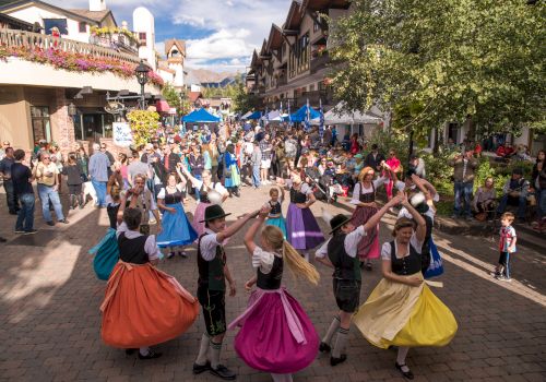 A festive street scene with people dancing in colorful skirts, vendors, and crowds lining a sunny cobblestone avenue&mdash;spirited celebration all around.