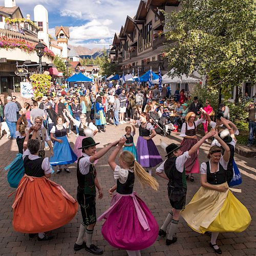 A festive street scene with people dancing in colorful skirts, vendors, and crowds lining a sunny cobblestone avenue&mdash;spirited celebration all around.