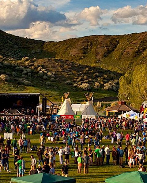 A large outdoor festival with a stage, tents, and a crowd on a grassy field set against green hills and a partly cloudy sky.