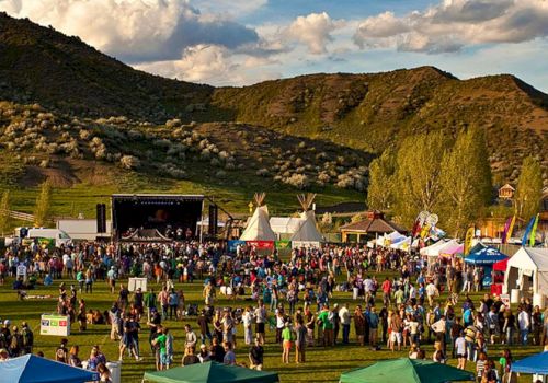 A large outdoor festival with a stage, tents, and a crowd on a grassy field set against green hills and a partly cloudy sky.