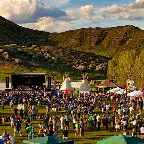 A large outdoor festival with a stage, tents, and a crowd on a grassy field set against green hills and a partly cloudy sky.