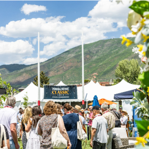 A lively outdoor festival with a crowd browsing stalls, tents, and a stage under a bright, sunny sky in a mountain setting.