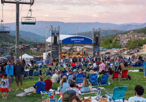 A large outdoor concert or festival in a grassy area with a stage, crowd, chairs, tents, and a ski lift in the background, under a clear sky.