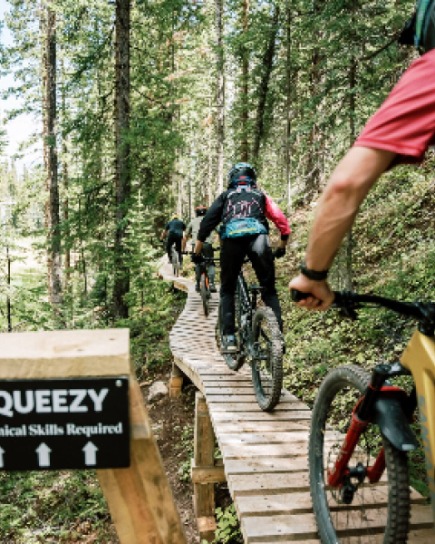 A group of mountain bikers rides along a wooden trail in a forest; a sign reads &ldquo;SQUEEZY&rdquo; and &ldquo;Technical Slopes Required&rdquo; at the front.