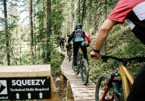 A group of mountain bikers rides along a wooden trail in a forest; a sign reads &ldquo;SQUEEZY&rdquo; and &ldquo;Technical Slopes Required&rdquo; at the front.