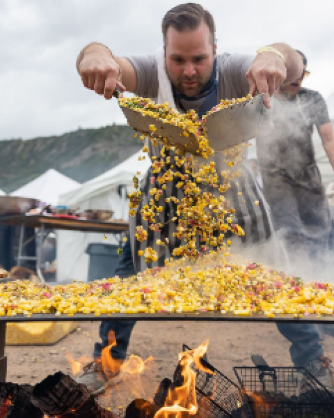 Two men grill a massive pan of stir-fry over an open flame, tossing corn, peppers, and vegetables into sizzling chaos at a street festival.