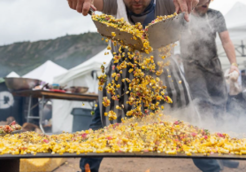 Two men grill a massive pan of stir-fry over an open flame, tossing corn, peppers, and vegetables into sizzling chaos at a street festival.