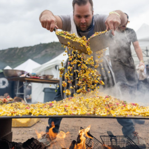 Two men grill a massive pan of stir-fry over an open flame, tossing corn, peppers, and vegetables into sizzling chaos at a street festival.