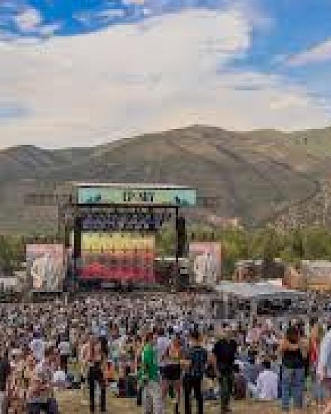 A large outdoor music festival with a stage, crowd, and mountains in the background, plus a gondola lift in the sky.