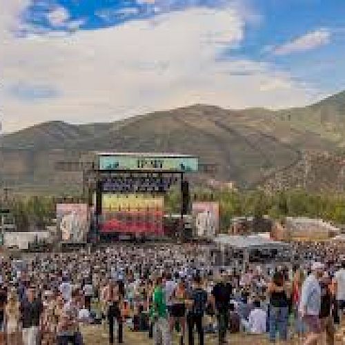 A large outdoor music festival with a stage, crowd, and mountains in the background, plus a gondola lift in the sky.