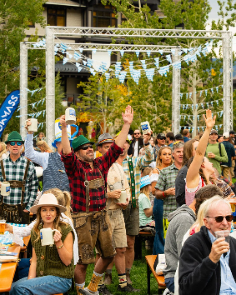 A lively outdoor festival with a crowd enjoying drinks, casual wear, tents, and a decorative arch under sunny trees, everyone smiling and cheering.