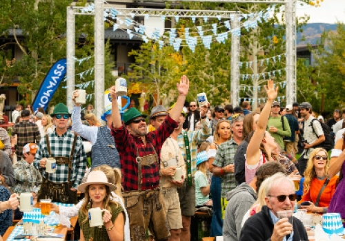 A lively outdoor festival with a crowd enjoying drinks, casual wear, tents, and a decorative arch under sunny trees, everyone smiling and cheering.