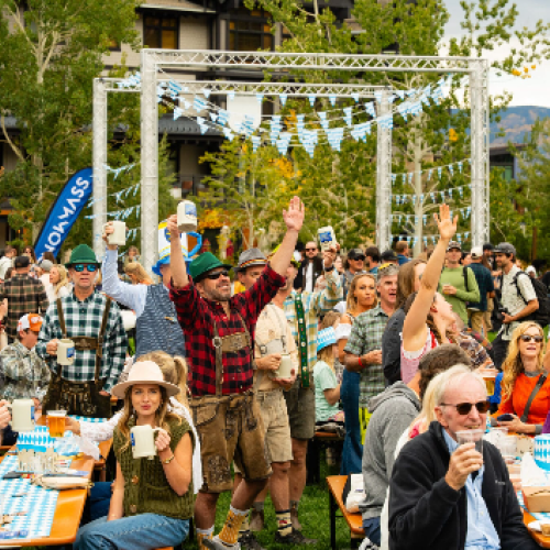 A lively outdoor festival with a crowd enjoying drinks, casual wear, tents, and a decorative arch under sunny trees, everyone smiling and cheering.