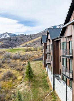 A row of wooden apartment buildings along a hillside with dry grass, rolling hills, and distant mountains under a partly cloudy sky.