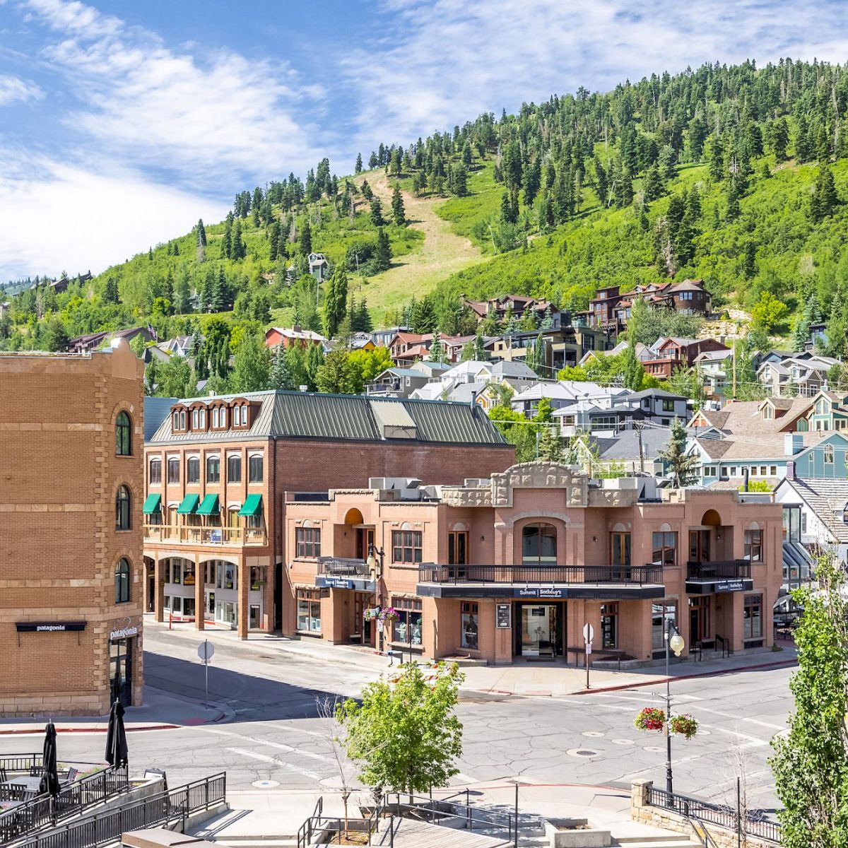 A small mountain town square with brick buildings, green hills, and a mix of shops; sunny day, trees, and clear blue sky.