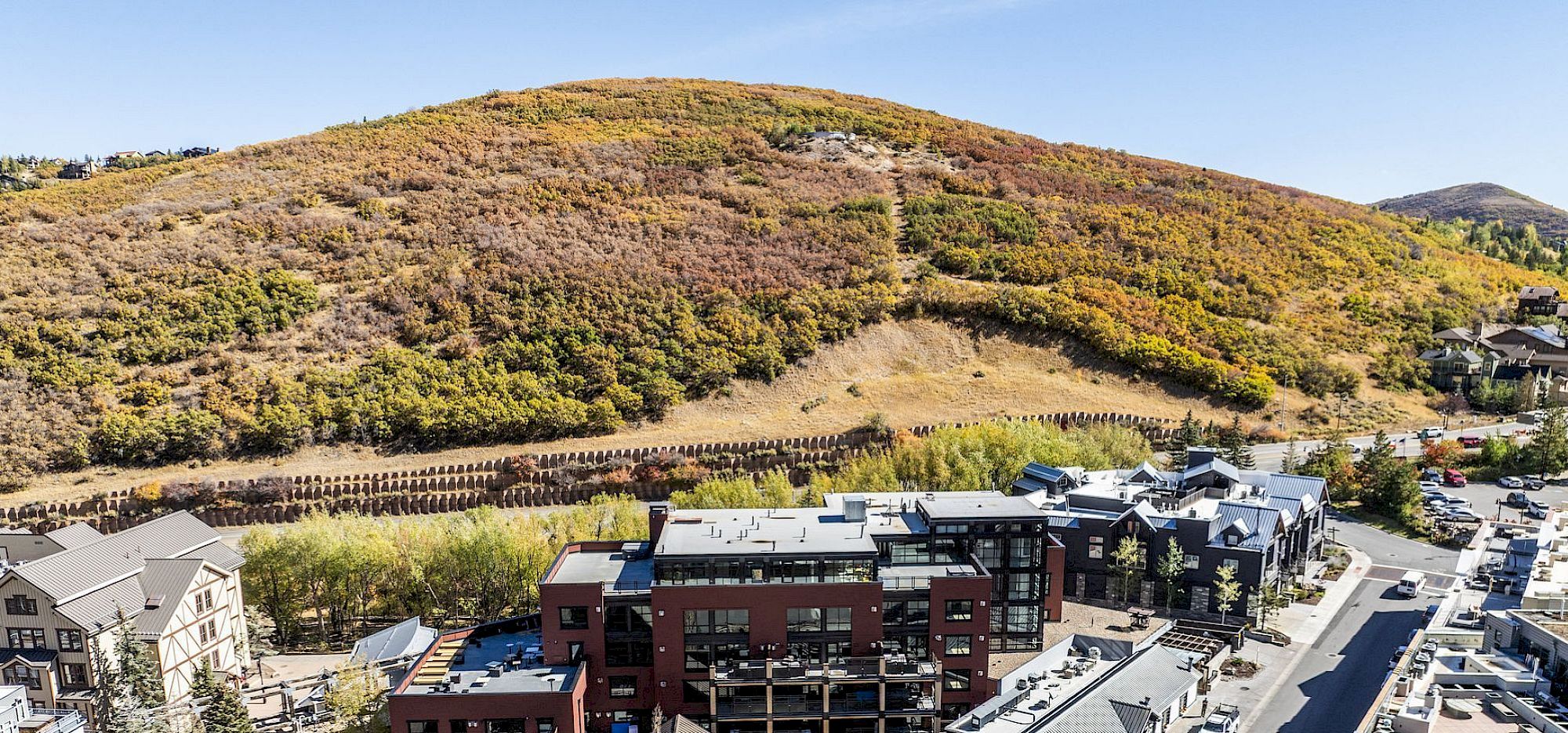Aerial view of a small town with mixed-use buildings along a winding street, set against a backdrop of rolling hill slopes and autumn foliage.