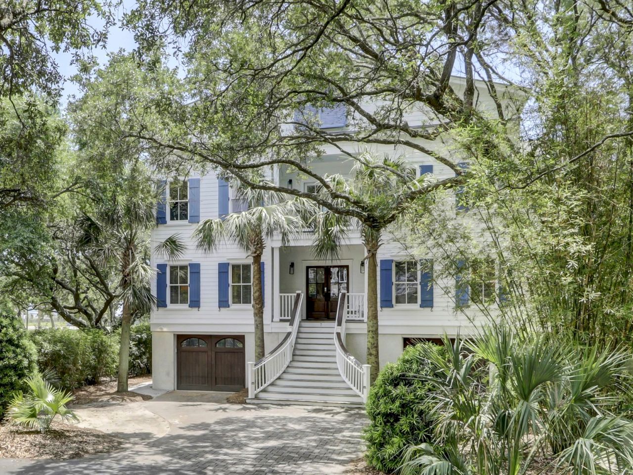 A large, two-story white house with blue shutters, surrounded by lush greenery and trees, featuring a double stairway at the entrance.