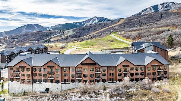 A row of brown apartment buildings at the base of a mountainous area with a winding road and snow-dusted hills in the background.