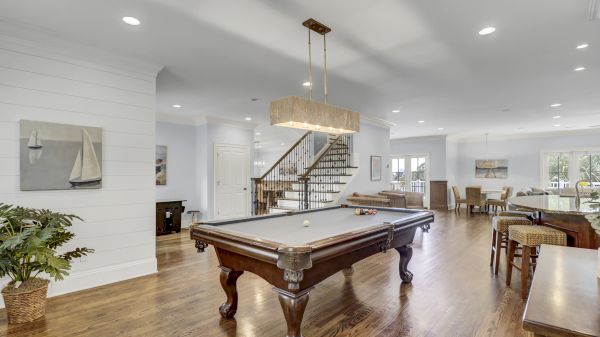 A spacious living area with a pool table, wooden floor, staircase, and modern dining setup, featuring soft lighting and decor.